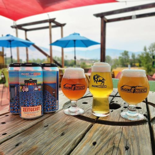 Two glasses of beer sit on a wooden table with a colorful can of "Zeitgeist" beer in the background and umbrellas overhead.