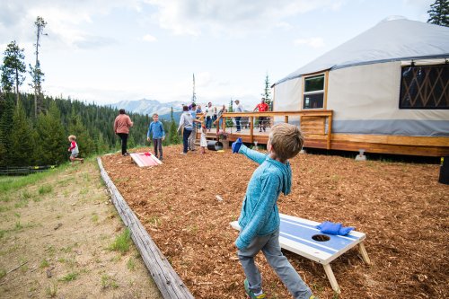 A child throws a bean bag towards a cornhole board near a yurt, with people socializing in the background and mountains beyond.