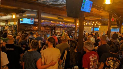 A crowded bar scene with people celebrating, wearing party hats, and enjoying drinks under warm lighting.