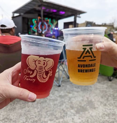 Two hands clink plastic cups of red drink and beer at an outdoor event, with a stage and audience in the background.