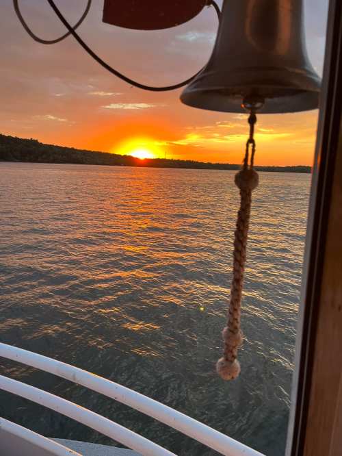 A sunset over calm water, viewed from a boat, with a bell hanging in the foreground.