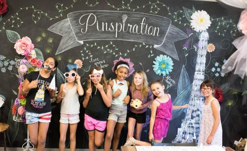 A group of seven girls poses playfully in front of a decorated chalkboard backdrop that says "Pinspiration."