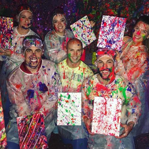 A group of six people in paint-splattered outfits hold colorful canvases, smiling in a vibrant, artistic setting.