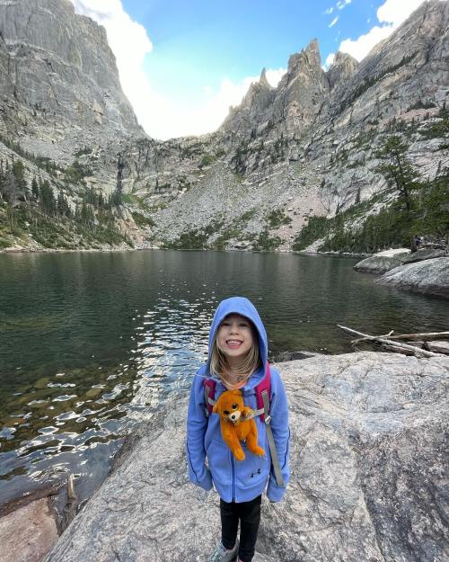 A smiling child in a blue hoodie stands by a lake, holding a stuffed animal, surrounded by mountains and trees.