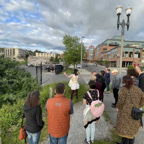 A group of people listens to a speaker in a park-like area near a city street, with buildings in the background.