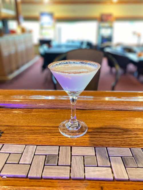 A cocktail in a martini glass with a salted rim, sitting on a wooden bar top. Background features a blurred restaurant setting.