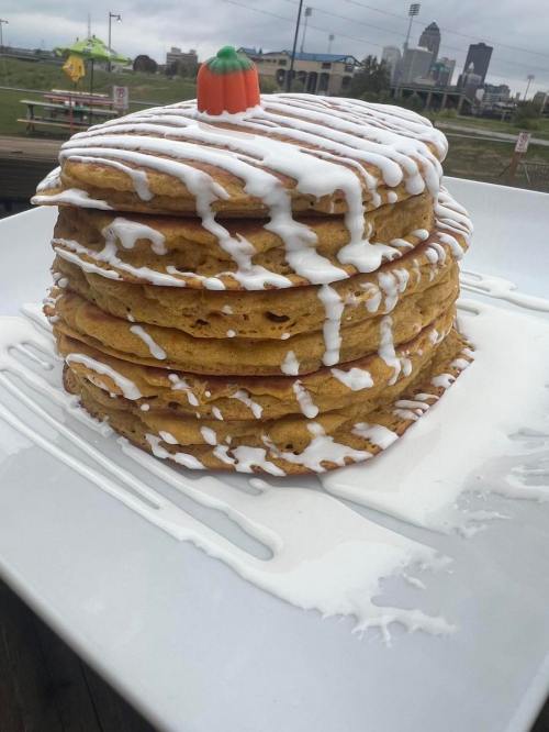 A stack of five large pancakes drizzled with white icing and topped with a small pumpkin decoration on a white plate.