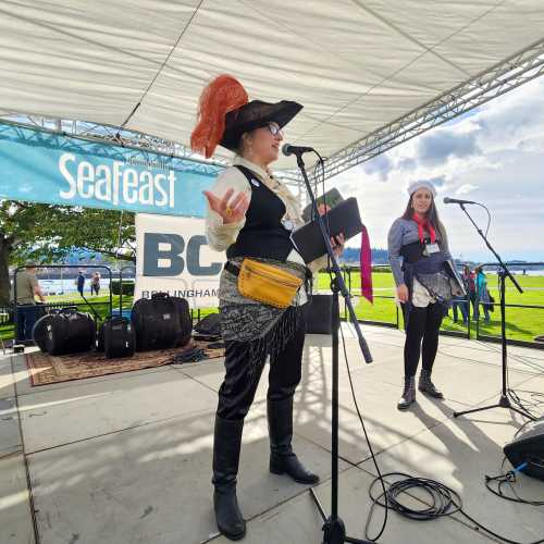 Two women on stage at a festival, one in a pirate costume with a feathered hat, speaking into a microphone.