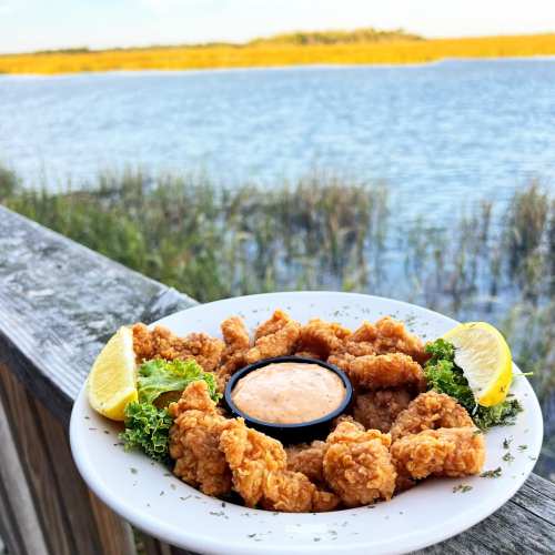 A plate of crispy fried shrimp with a dipping sauce, garnished with lemon and lettuce, by a scenic water view.