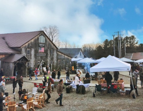 A bustling outdoor market scene with tents, people shopping, and a rustic barn in the background under a blue sky.