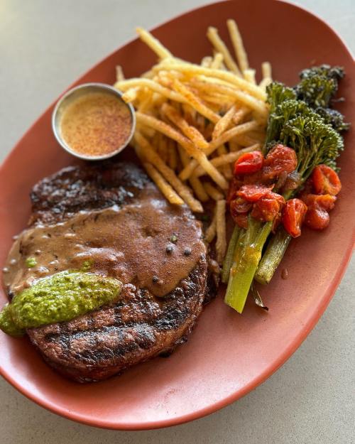 A plate featuring a grilled steak with sauce, crispy fries, roasted tomatoes, and broccolini on a red platter.