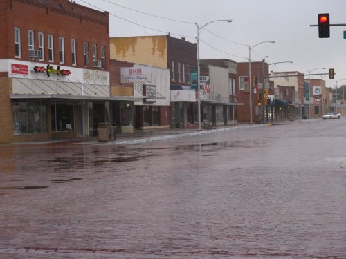 A quiet, rainy street scene with empty storefronts and wet cobblestones, under a gray sky. Traffic lights visible.