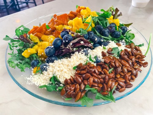 A colorful salad with greens, mango, blueberries, roasted pumpkin seeds, and crumbled cheese on a glass plate.