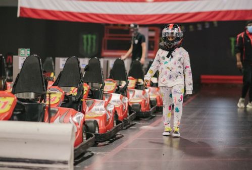 A child in a helmet walks past go-karts in a racing venue, wearing colorful pajamas and a focused expression.
