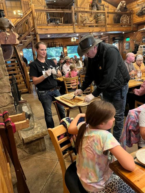 A restaurant scene with a man serving food while a woman assists, and a child watches at a wooden table.
