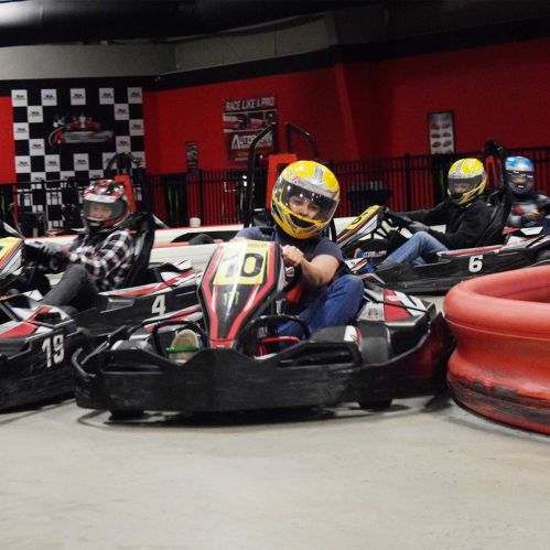 A group of go-karts racing on an indoor track, with drivers wearing helmets and racing gear.