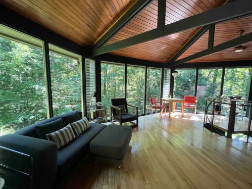 A modern living space with large windows, wooden ceiling, and minimalist furniture overlooking a forested area.