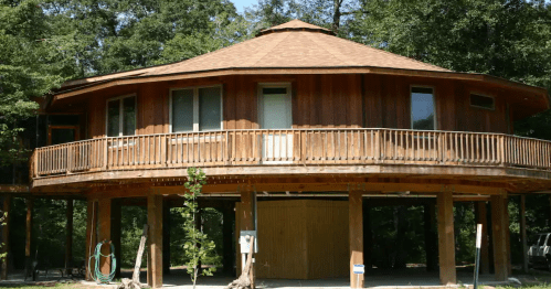 A unique, circular wooden home on tall pillars with a wide, curved balcony and shingled roof.