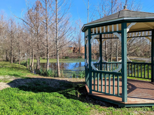A gazebo in a park surrounded by trees and a pond under a clear blue sky.