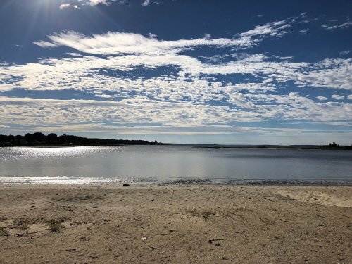 A serene beach scene with soft sand, calm water, and a sky filled with fluffy clouds and sunlight.