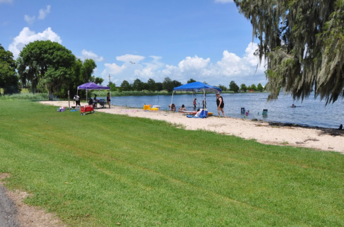 A sunny lakeside scene with people under blue tents, enjoying a sandy beach and green grass.