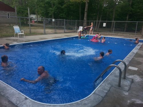 A lively outdoor pool scene with people swimming, lounging, and children playing in the water on a sunny day.