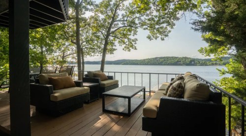 A cozy outdoor seating area with plush sofas and a coffee table, overlooking a serene lake surrounded by trees.