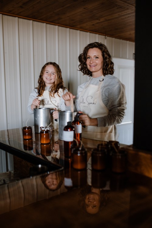 A woman and a girl in aprons smile while mixing ingredients in a kitchen, with amber jars on the counter.