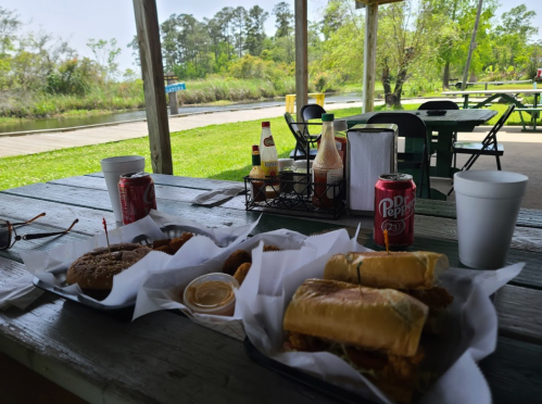 A picnic table with two trays of food, drinks, and condiments, set in a scenic outdoor area by a waterway.