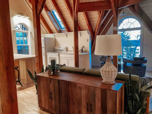 Cozy interior of a cabin with wooden beams, a lamp, and a view of nature through large windows.