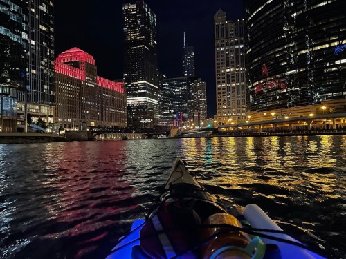 A kayak on a river at night, surrounded by illuminated skyscrapers and reflections on the water.