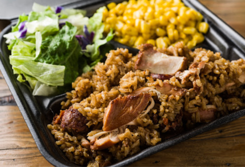 A black takeout tray with seasoned rice, grilled chicken, corn, and a side salad of lettuce and purple cabbage.