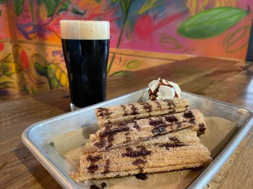 A plate of churros drizzled with chocolate sauce, served with a scoop of ice cream, alongside a glass of dark beer.