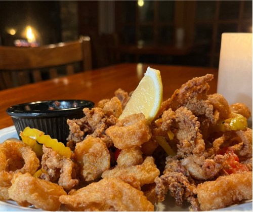 A plate of fried seafood with lemon and peppers, served with a side of dipping sauce. Warm, cozy restaurant setting.
