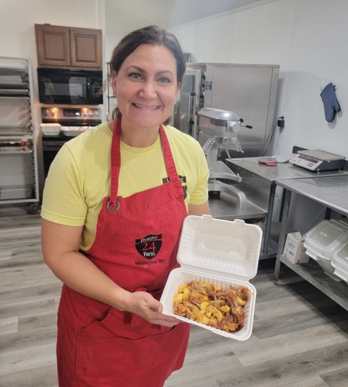 A smiling woman in a yellow shirt and red apron holds a takeout container filled with food in a kitchen setting.