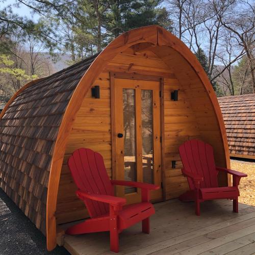 A cozy wooden cabin with a curved roof, featuring two red chairs on the porch and surrounded by trees.