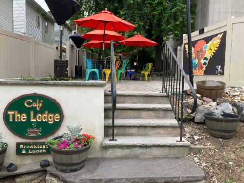 A cozy café entrance with stairs, colorful chairs, and red umbrellas in a garden setting. Flowers and decor enhance the scene.