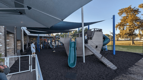 A playground with a wooden structure, slides, and shaded areas, surrounded by trees and a clear blue sky.
