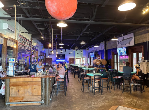 A lively bar interior with wooden decor, patrons seated at tables, and a large red planet hanging from the ceiling.