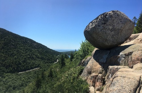 A large boulder perched on a rocky ledge overlooking a lush green valley and distant water under a clear blue sky.