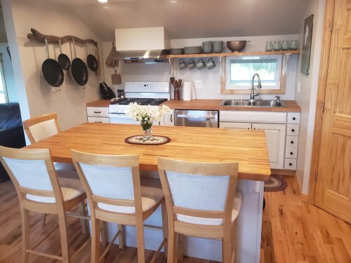 A cozy kitchen with a wooden island, four chairs, pots hanging, and a window above the sink. Bright and inviting space.