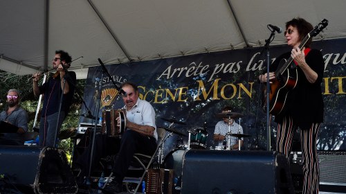 A band performs on stage under a tent, featuring musicians with various instruments and a backdrop with text.
