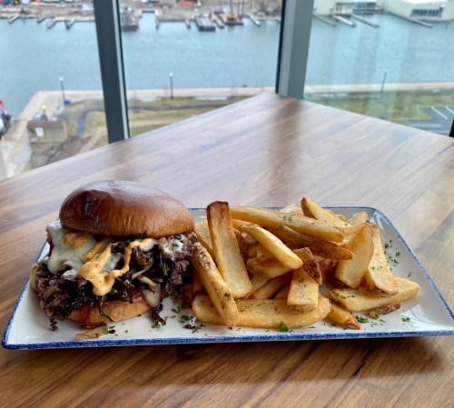 A juicy burger with melted cheese and sauce, served with crispy fries, on a wooden table with a waterfront view.