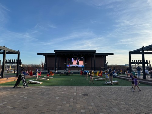 A lively outdoor space with a large screen, people playing games, and a grassy area under a clear blue sky.