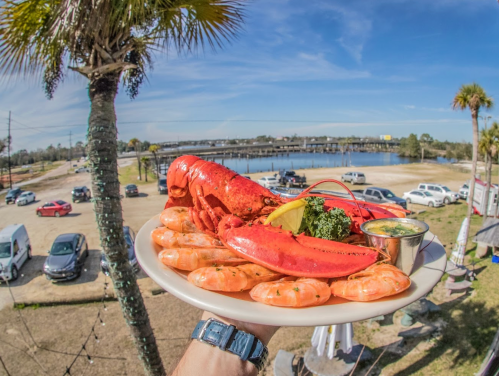 A plate of lobster and shrimp garnished with lemon and sauce, held against a sunny waterfront backdrop.