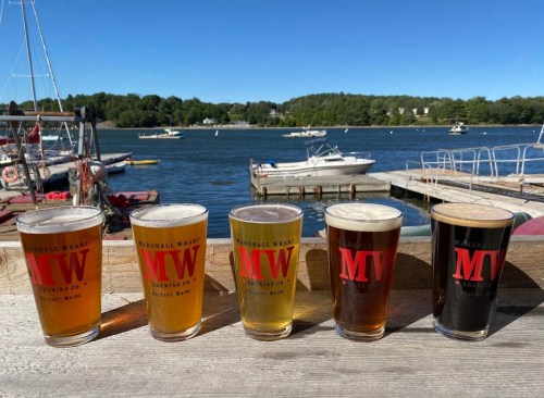 Four glasses of beer in front of a scenic waterfront with boats, featuring the logo "MTW" on each glass.