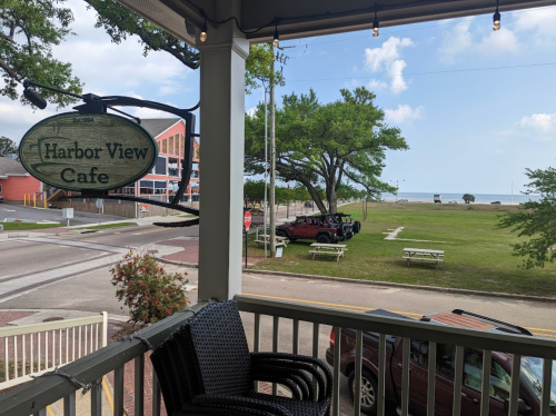 View from a cafe porch overlooking a grassy area, parked vehicle, and distant shoreline under a blue sky.