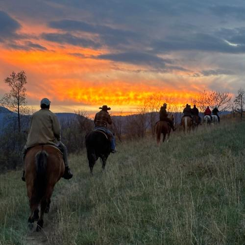 Cowboys riding horses along a grassy path at sunset, with vibrant orange and purple skies in the background.