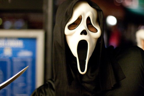 A person in a black hooded costume with a white ghost face mask, holding a knife, set against a blurred background.
