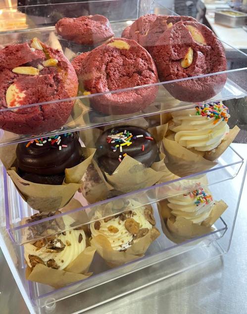 A clear display case filled with colorful cupcakes and red velvet cookies, topped with sprinkles and nuts.